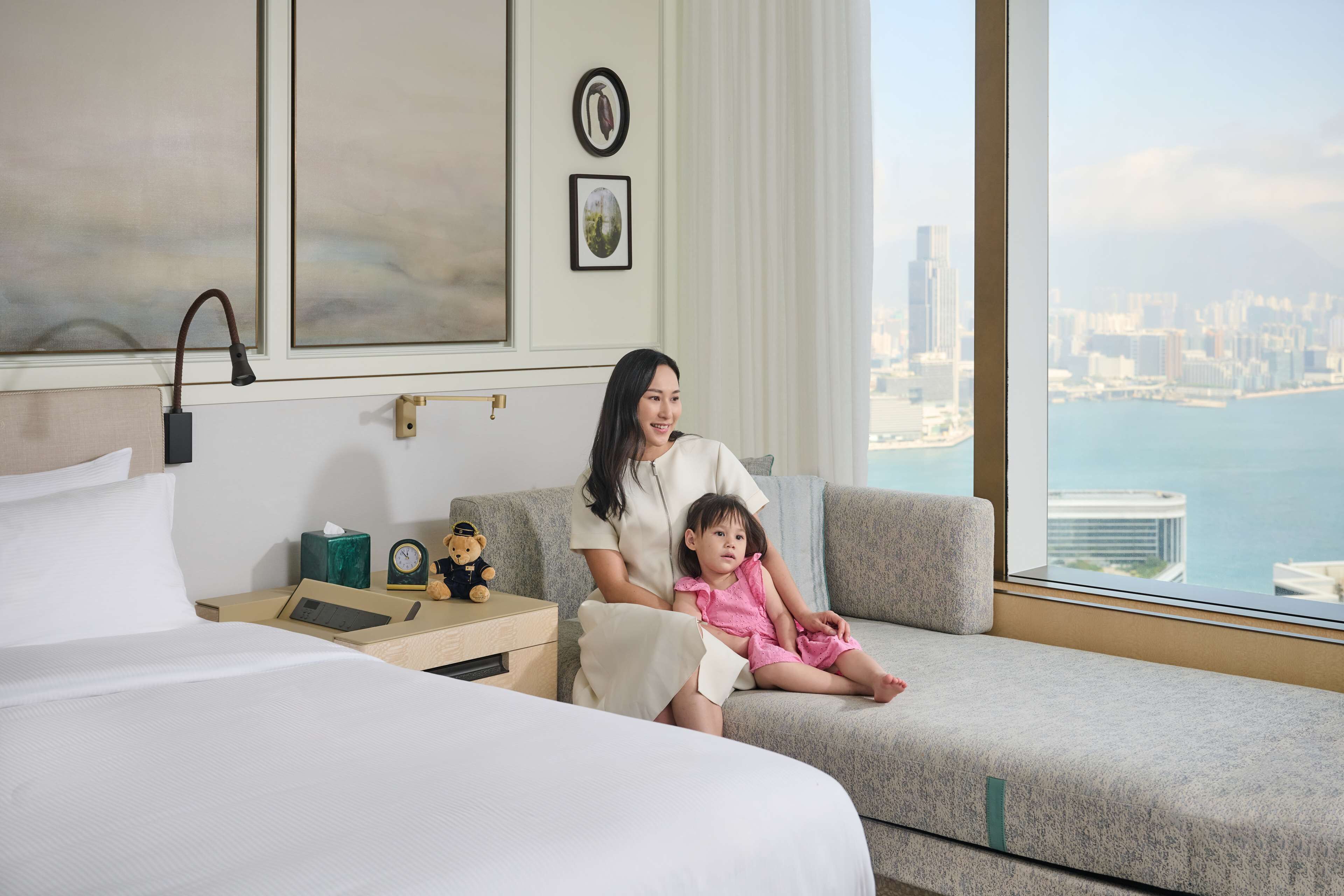 A hotel room with a city view, a bed, a sofa, and a nightstand with a clock and a teddy bear.