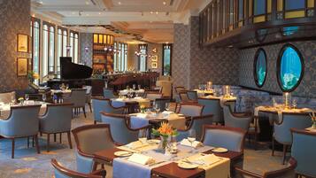 A spacious dining area with multiple tables and chairs, a piano, and large windows providing natural light.