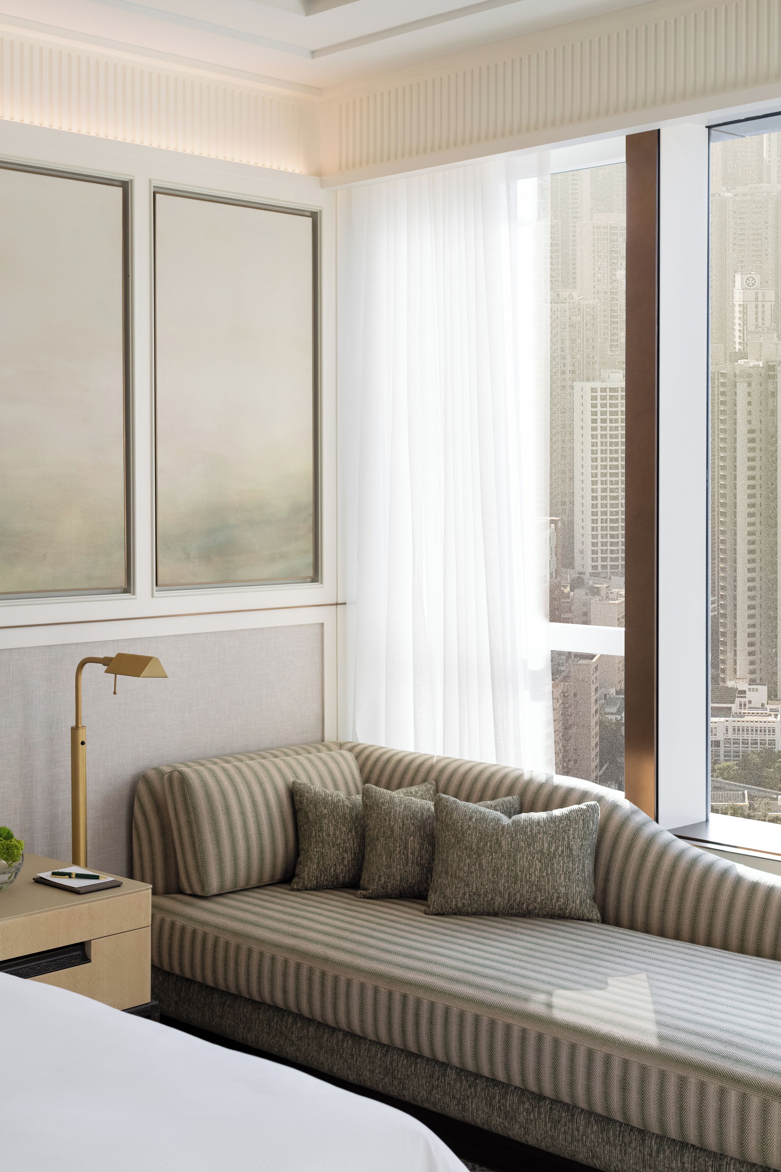 Una camera d'albergo moderna con un'ampia finestra, un divano a righe, un comodino con lampada e vista sulla città.