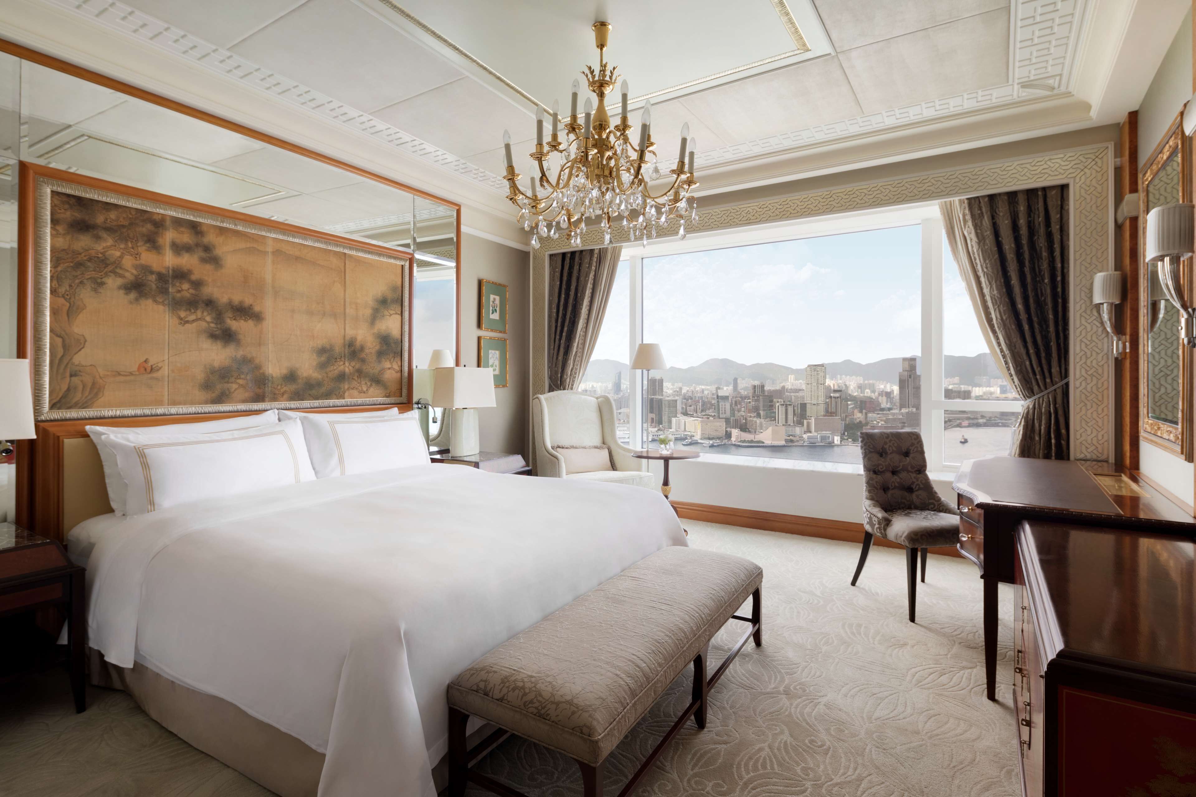 A spacious hotel room with a large bed, a bench, a desk, and a view of the city.