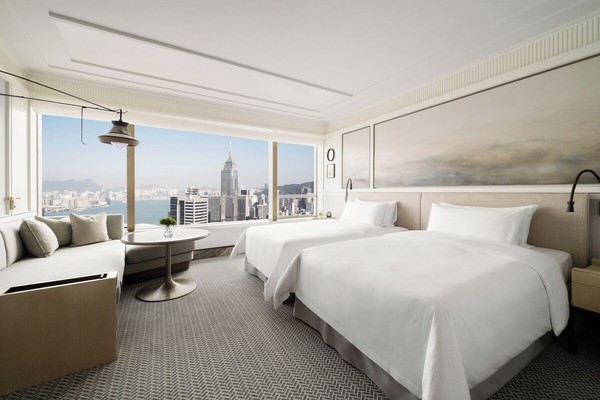 A hotel room with two beds, a sofa, a round table, and a large window with a city view.