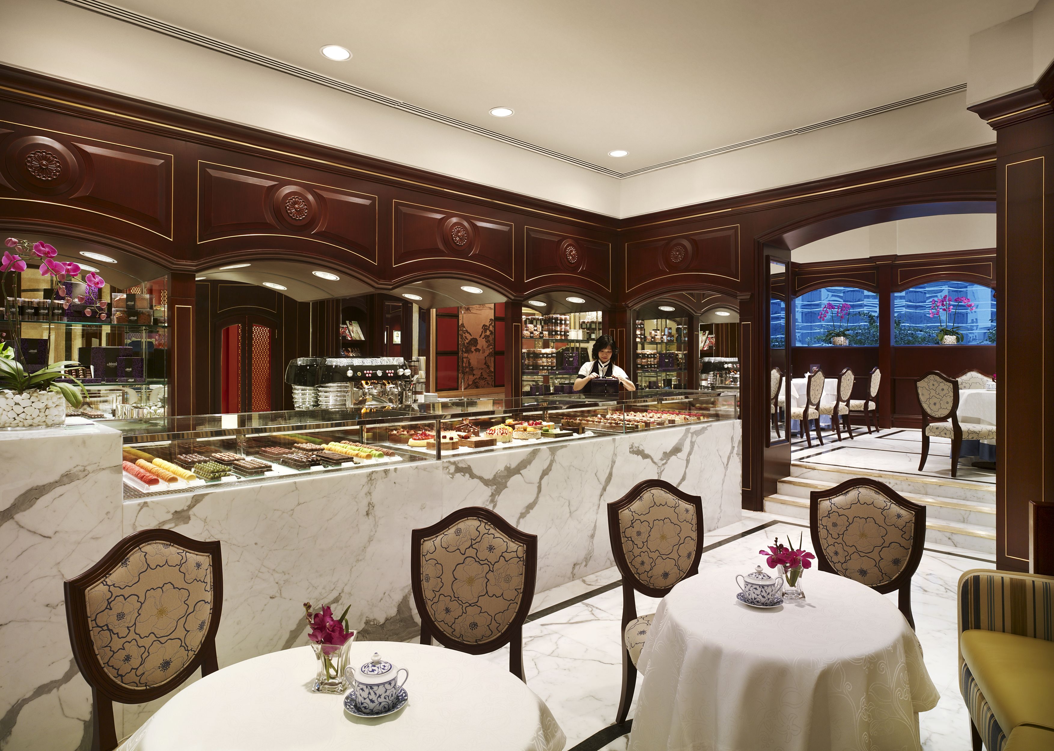 A luxurious hotel lobby with marble flooring, ornate wood paneling, a central dessert display, and elegantly set dining tables with chairs.