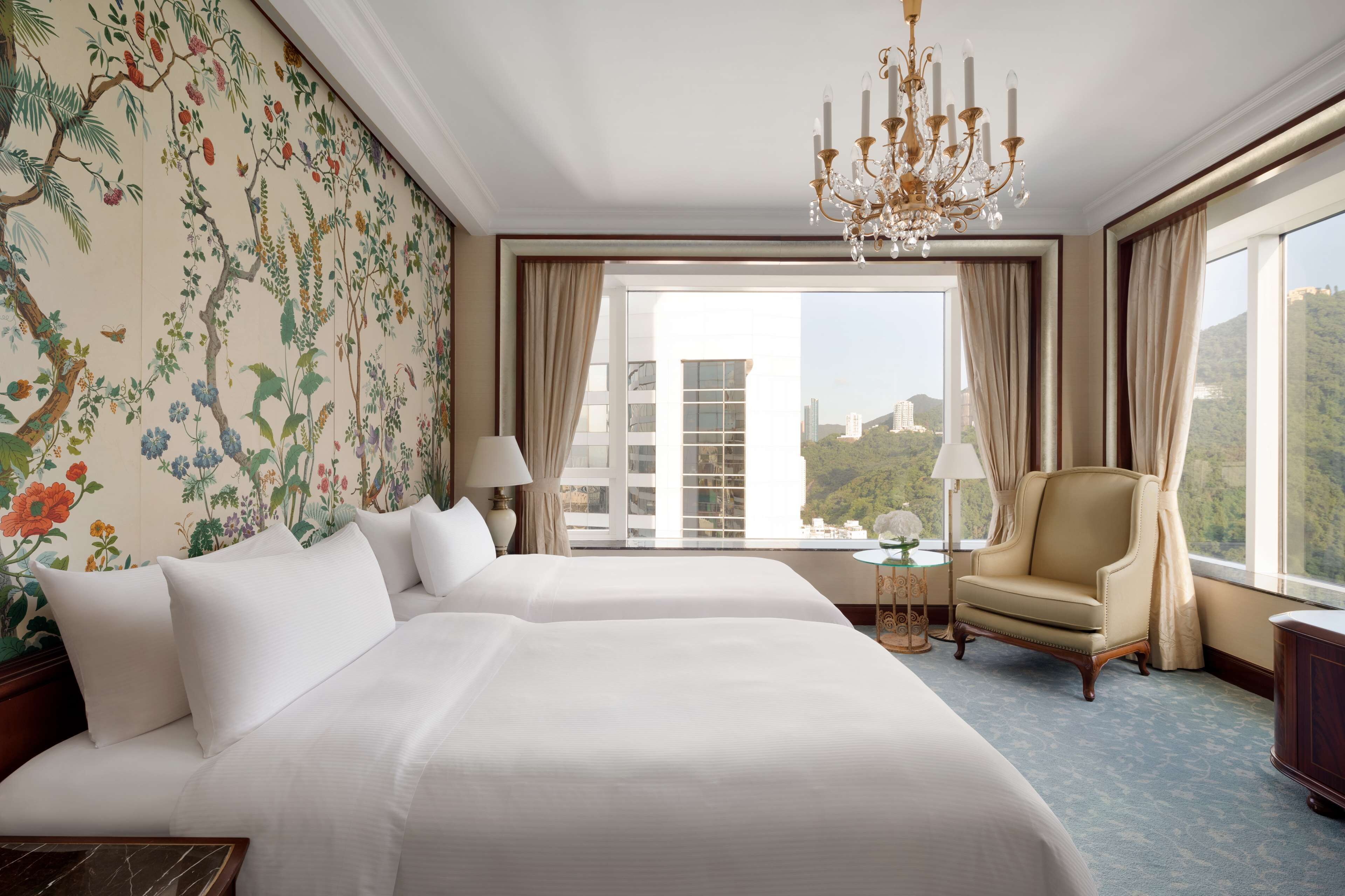 A hotel room with two beds, a chandelier, a chair, and a view of a mountainous landscape.