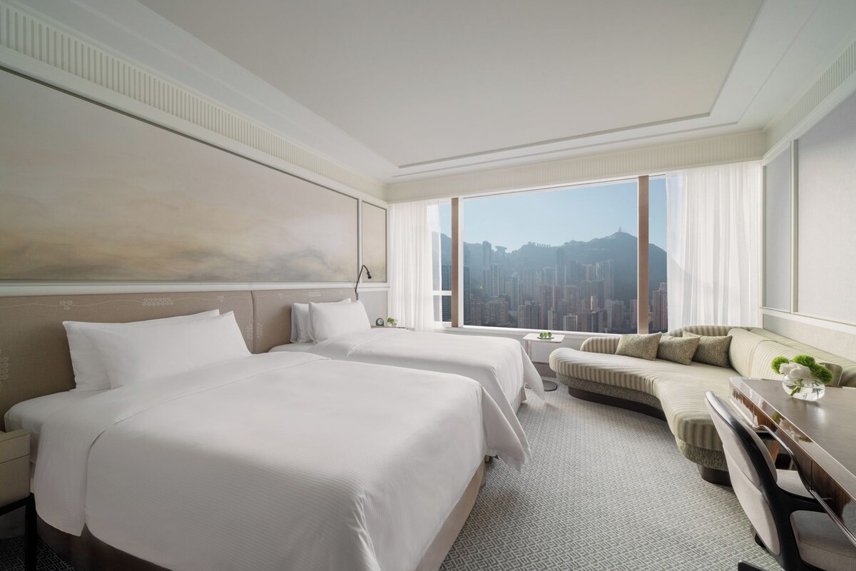 A hotel room with two beds, a sofa, a desk, and a large window with a city view.