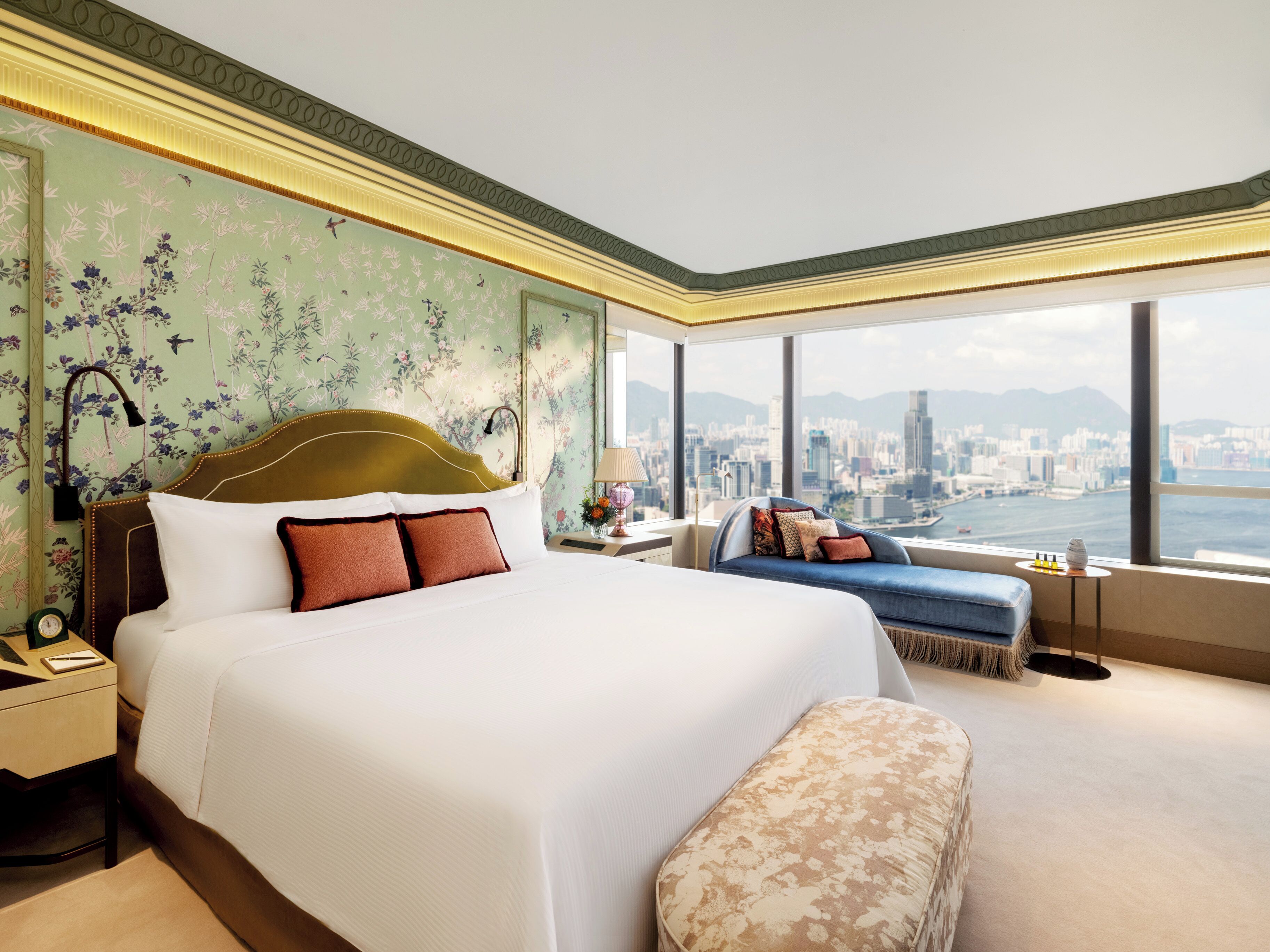 Habitación de hotel con una cama grande, zona de estar y vistas a un paisaje urbano a través de amplios ventanales.