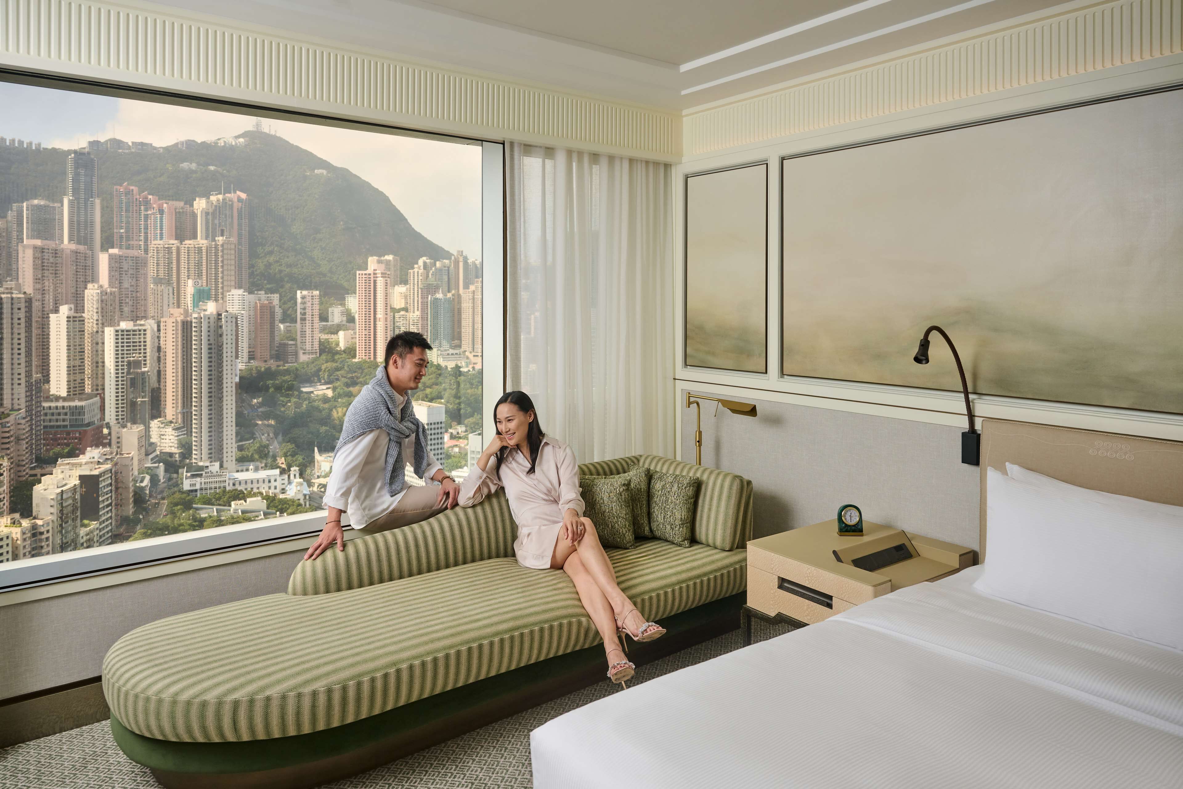 A hotel room with a large window offering a city view, a green sofa, a bed, and a nightstand with a lamp.