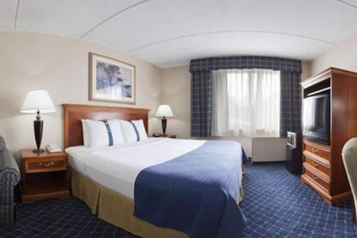 A hotel room with a large bed, two bedside tables with lamps, a television, a seating area with a chair, and a window with curtains.