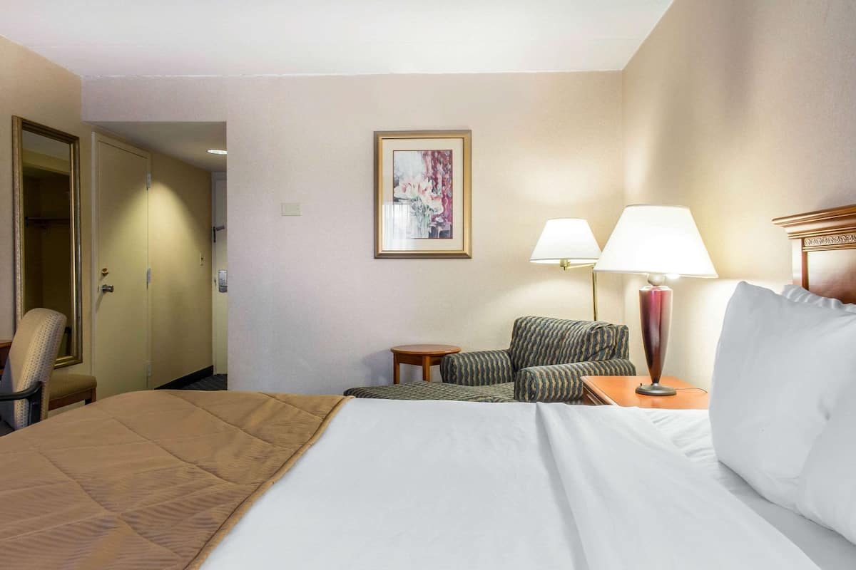 A hotel room with a bed, two chairs, a small table, a lamp, and a painting on the wall.