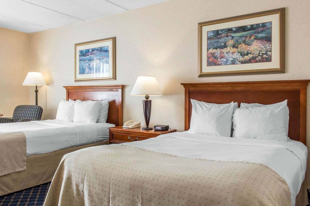 A hotel room with two beds, a chair, a nightstand with a lamp, and two framed pictures on the wall.