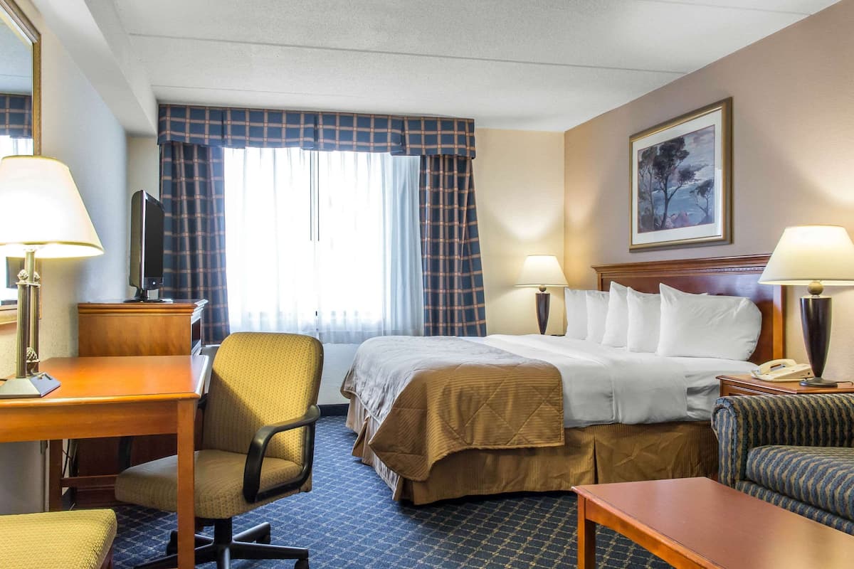 A hotel room with a bed, two bedside tables with lamps, a desk with a chair, a sofa, and a large window with curtains.
