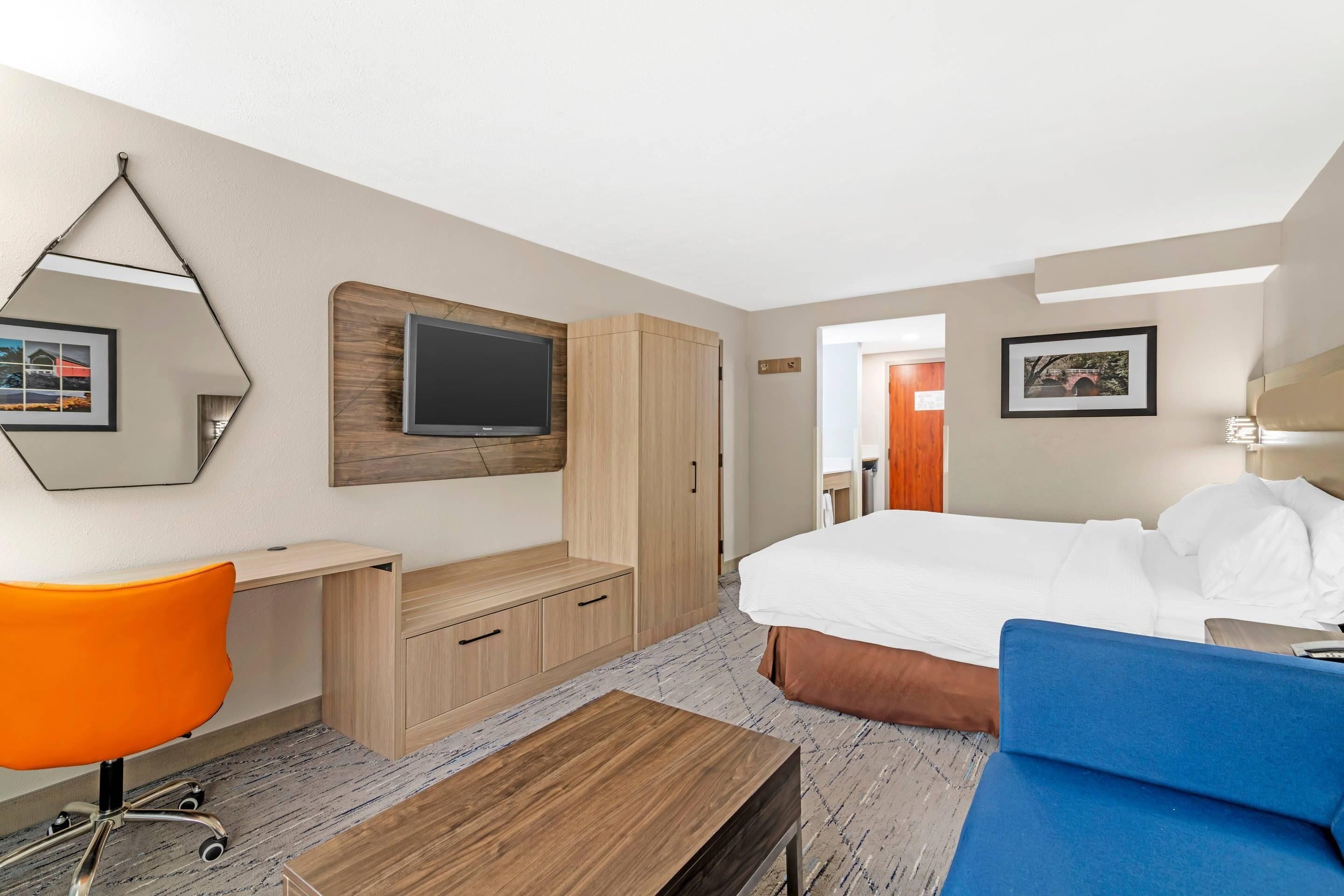 A hotel room with a bed, a desk, an orange chair, a TV, and a wardrobe.