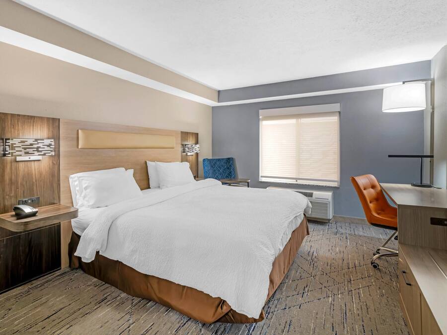 Ein Hotelzimmer mit einem großen Bett, einem Schreibtisch mit Stuhl, einem Fenster mit Jalousien und einem an der Wand montierten Fernseher.