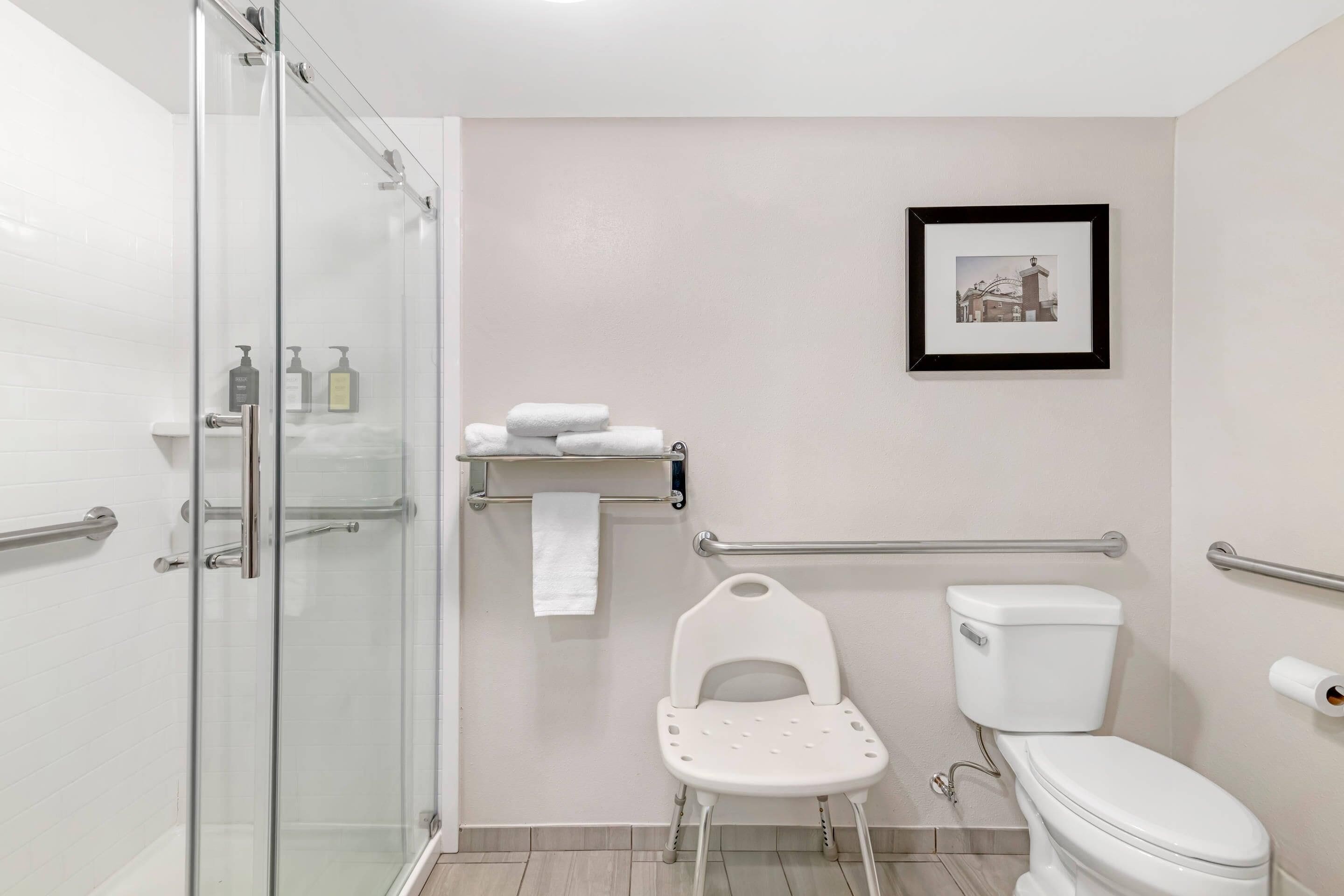 A bathroom with a glass shower enclosure, a white toilet, a white chair, and a towel rack.
