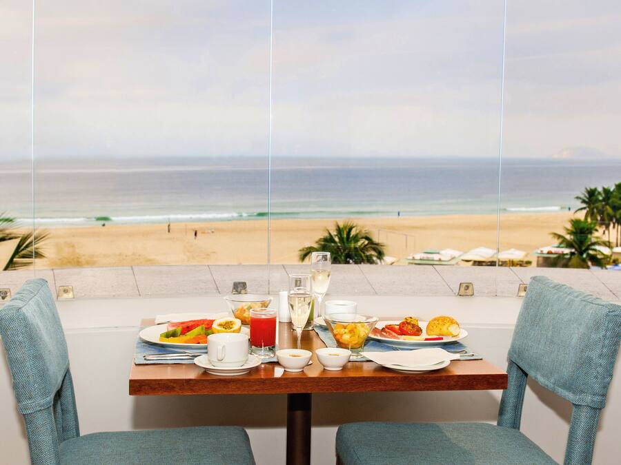 Una mesa de comedor con alimentos para el desayuno y un vaso de jugo, con vista a una playa con palmeras y vistas al océano.