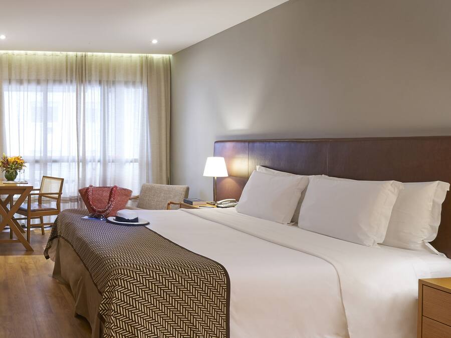 Habitación de hotel con una cama grande, una mesita de noche de madera, un escritorio con una silla y una ventana con cortinas translúcidas.