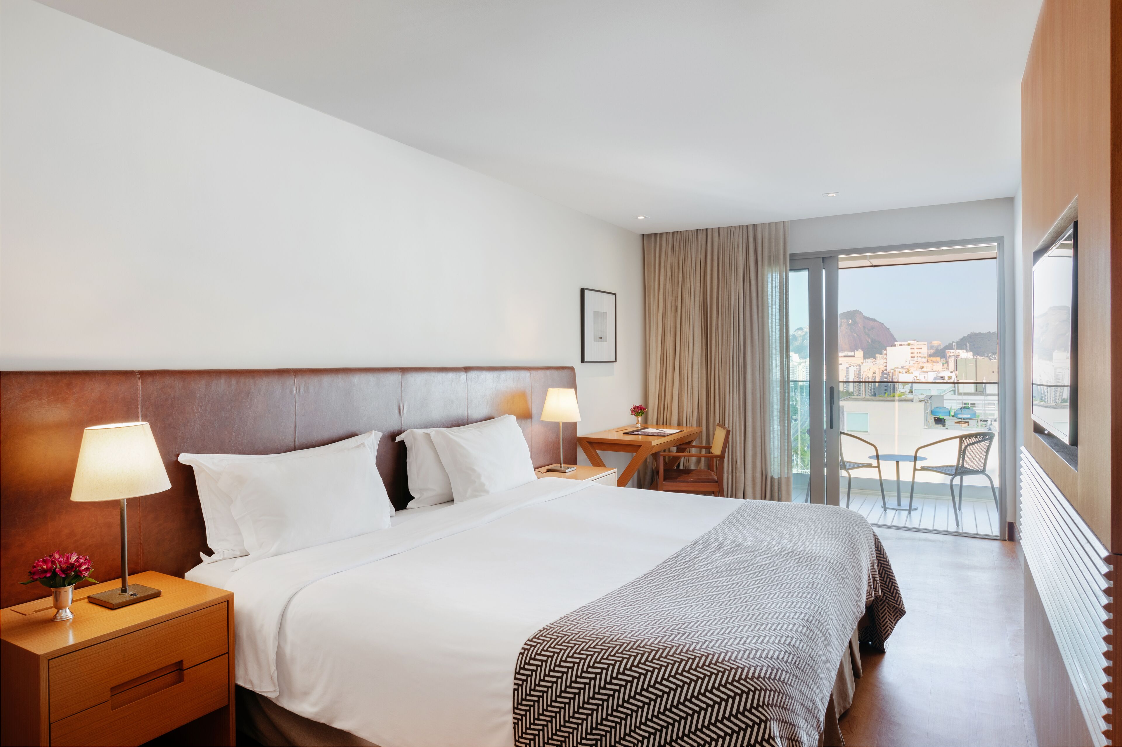 A hotel room with a large bed, two bedside tables with lamps, a desk with a chair, and a balcony with a view of the city.