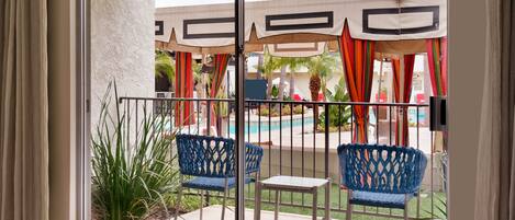A balcony with two blue chairs, a small table, and a view of a pool area with umbrellas and plants.