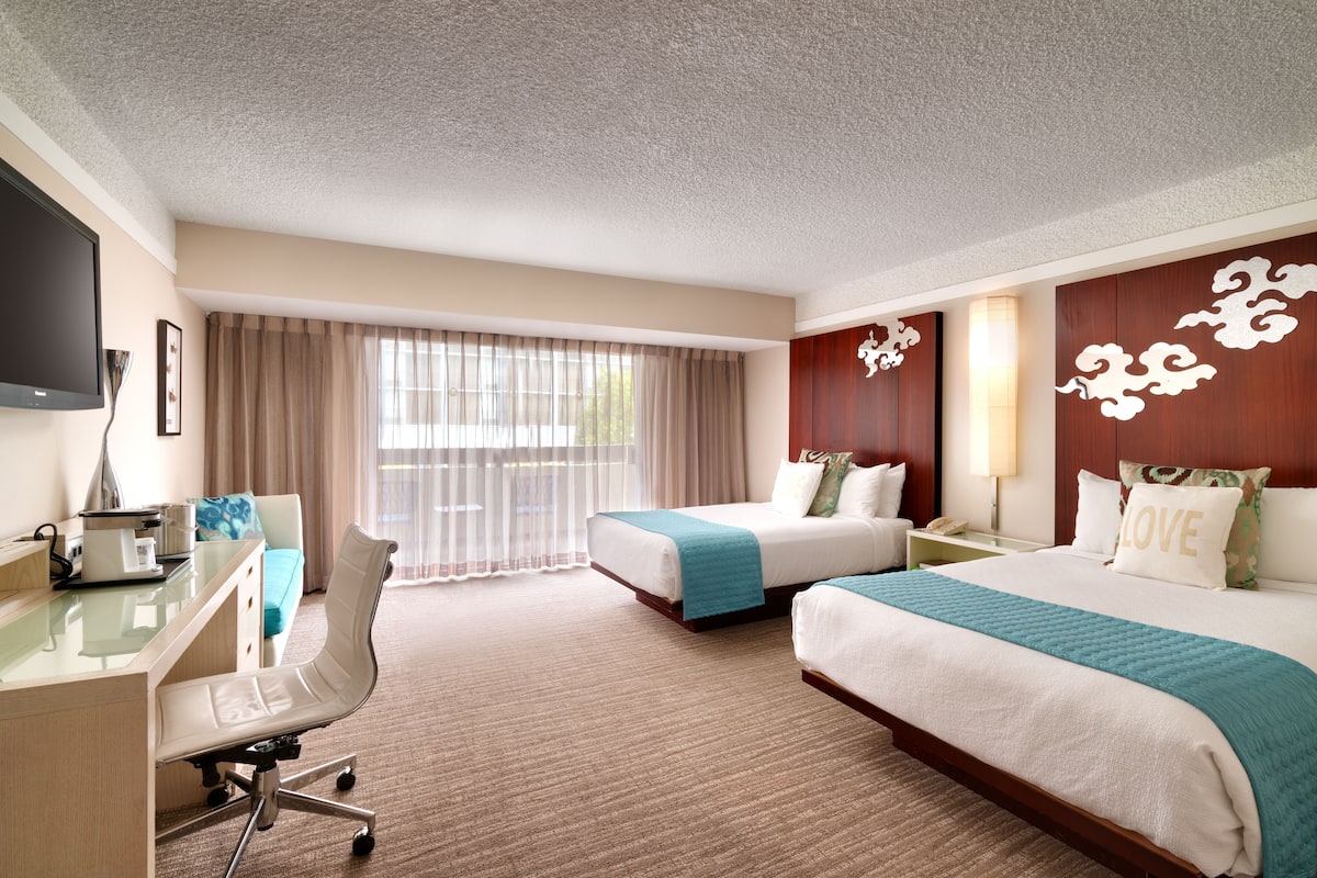 A hotel room with two beds, a desk, a chair, a TV, and a window with curtains.