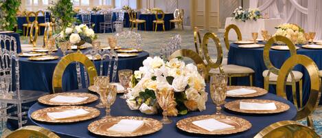 A banquet hall with round tables set for a formal event, featuring gold and white tableware, clear glassware, and floral centerpieces.