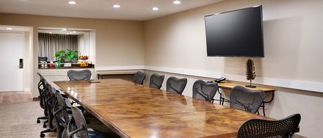 Una sala de conferencias con una mesa larga, sillas, un televisor de pantalla plana y una planta al fondo.