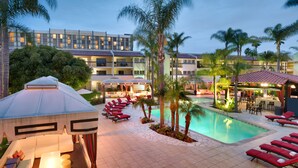 A hotel pool area with sun loungers, a covered seating area, and a building with multiple balconies. - Atrium Hotel at Orange County Airport (Irvine)