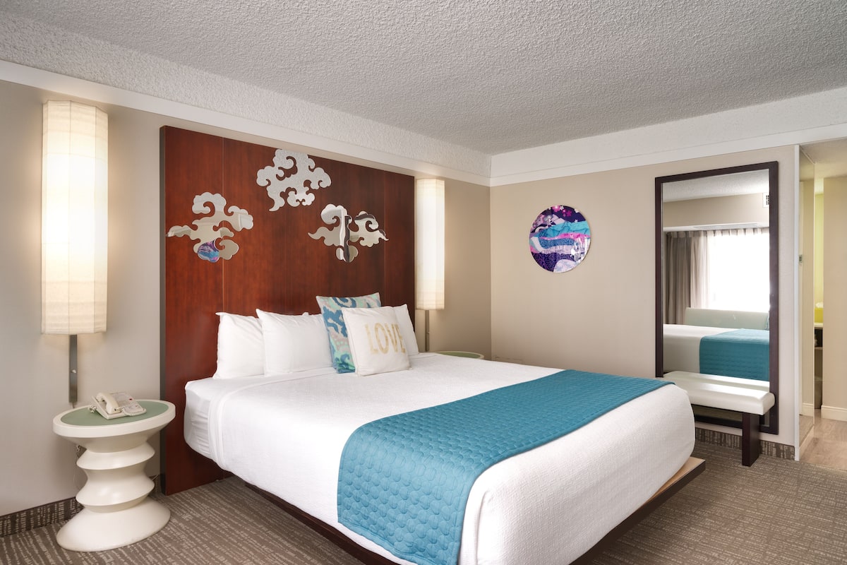 A hotel room with a large bed, a bedside table with a lamp, a wall clock, and a round mirror.