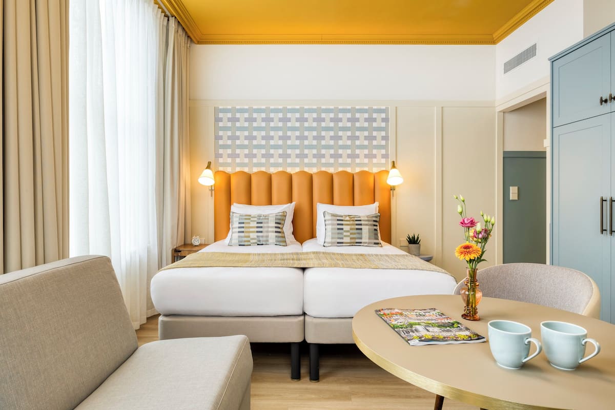 Una camera d'albergo con un letto matrimoniale, due comodini con lampade, un tavolino rotondo con due tazze e un vaso con fiori.
