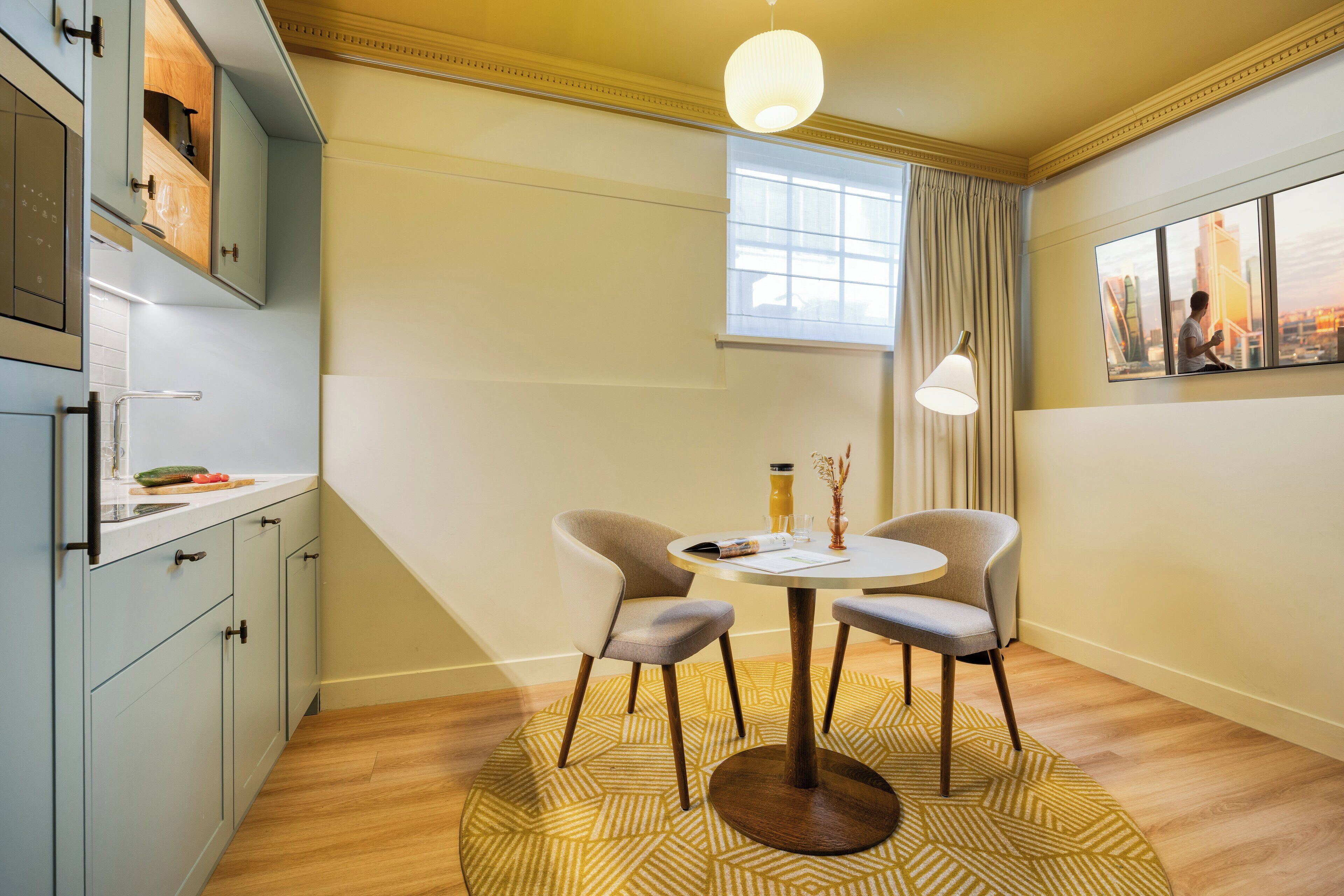 Une cuisine moderne dotée d’un four encastré, d’un coin repas avec une table ronde et deux chaises, et d’une grande fenêtre avec des rideaux.