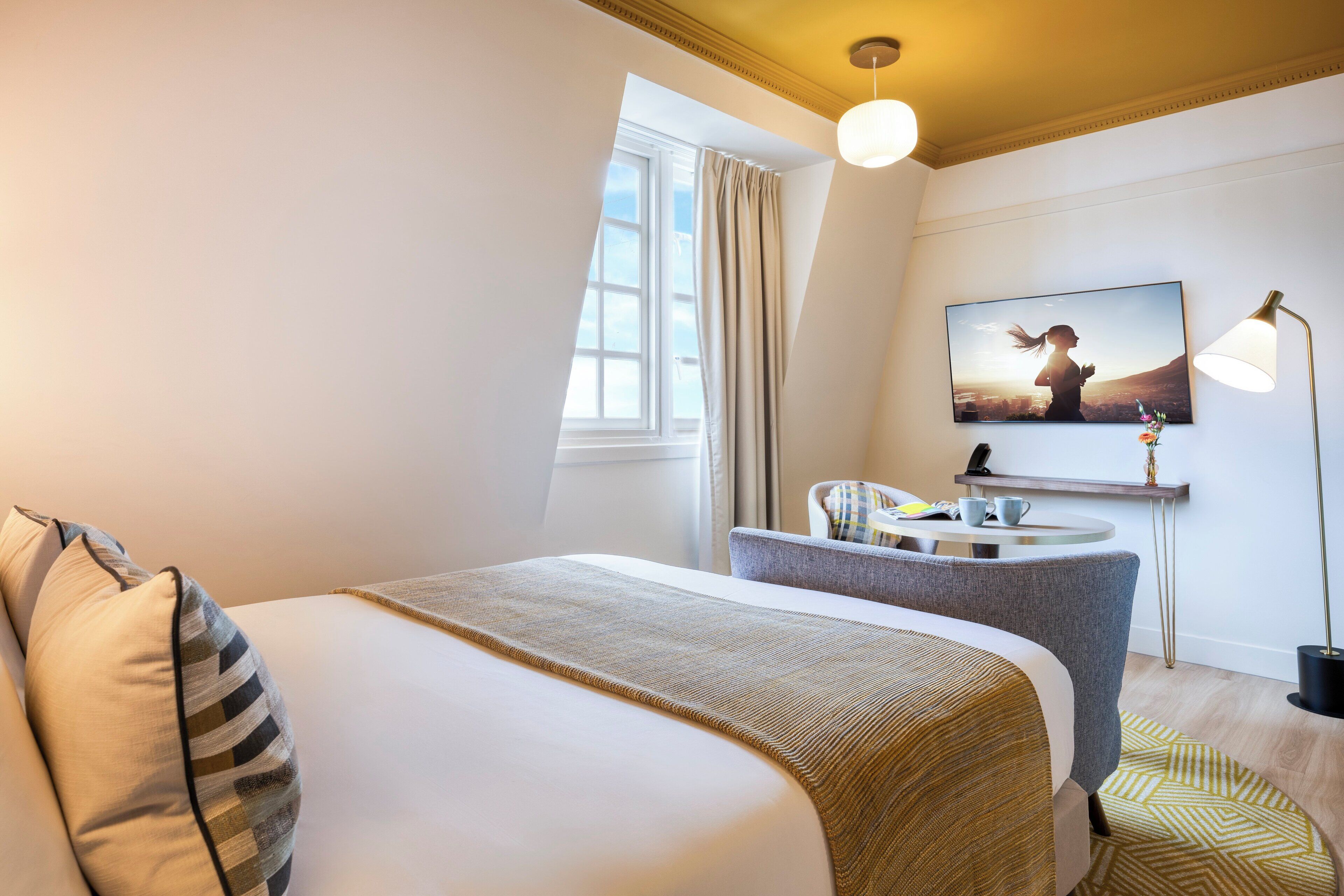 Habitación de hotel con cama, escritorio, silla, ventana, un cuadro y una lámpara.