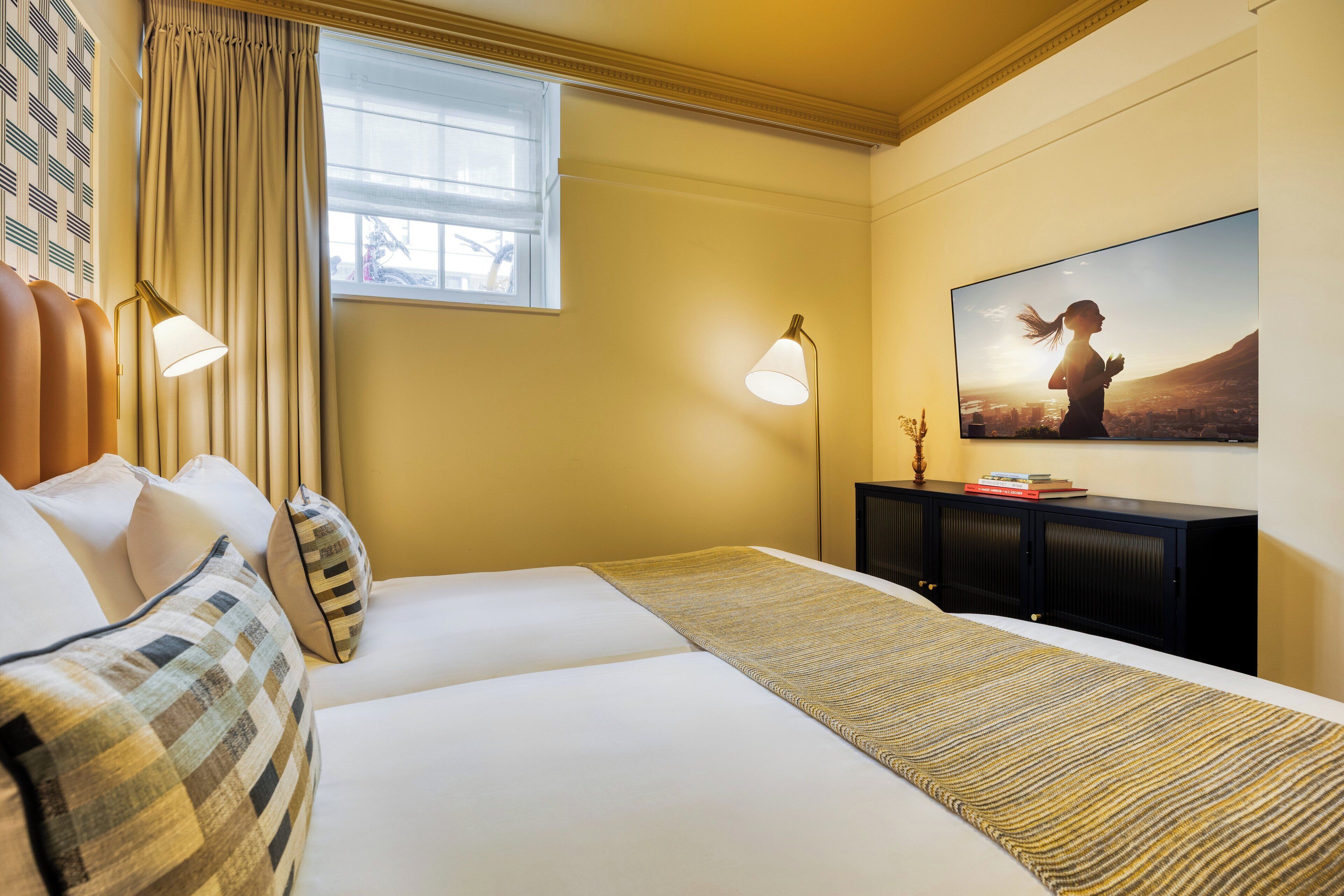 A hotel room with a bed, a window with curtains, a wall-mounted TV, and a bedside table with a lamp.