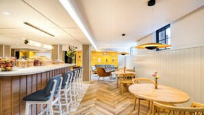 Zona de bar moderna con suelo de parqué, taburetes blancos y una barra que exhibe diferentes productos alimenticios.