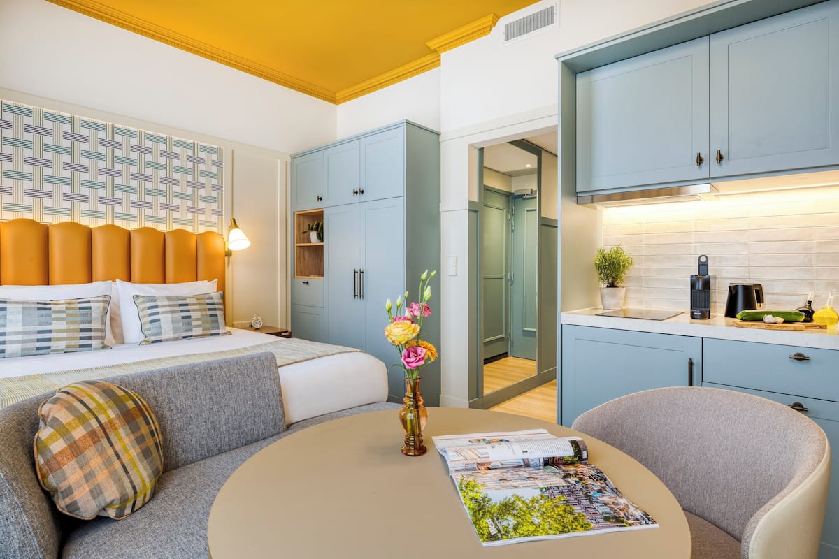 Una camera d'albergo moderna con un letto rifatto, un divano, un tavolino con un vaso di fiori e un angolo cottura con pensili e un piano di lavoro.
