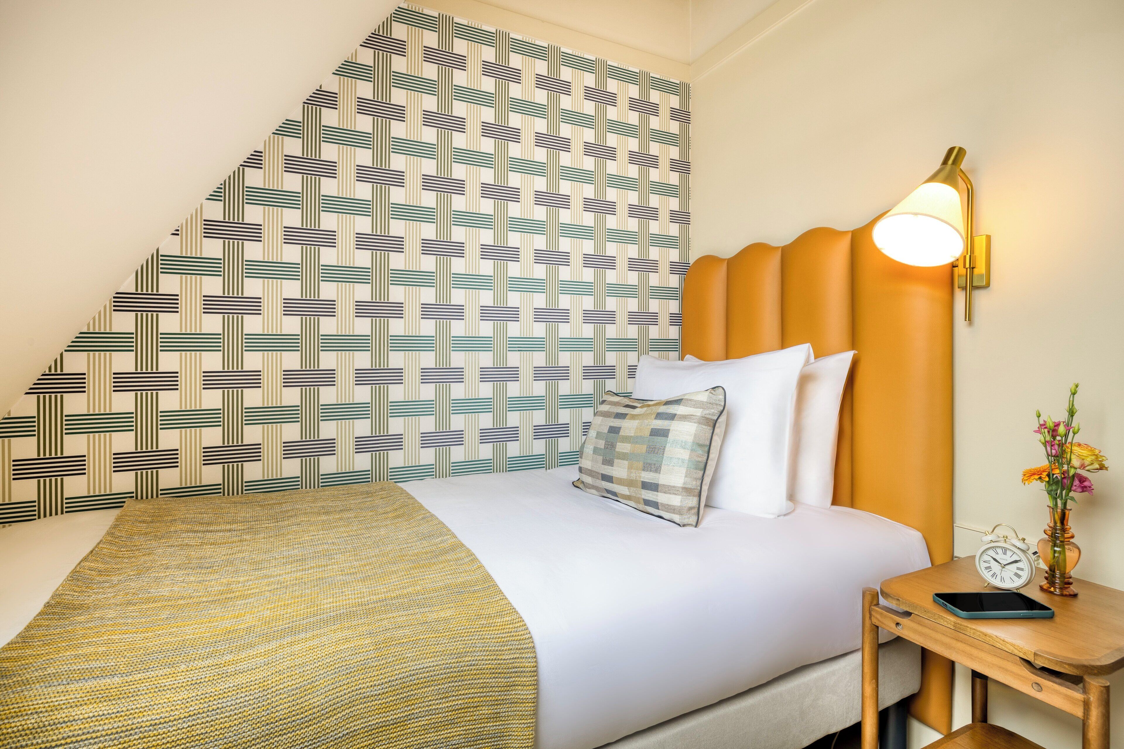 A hotel room with a patterned wall, a bed with white bedding, a yellow throw, a bedside table with a lamp, a clock, and a vase with flowers.