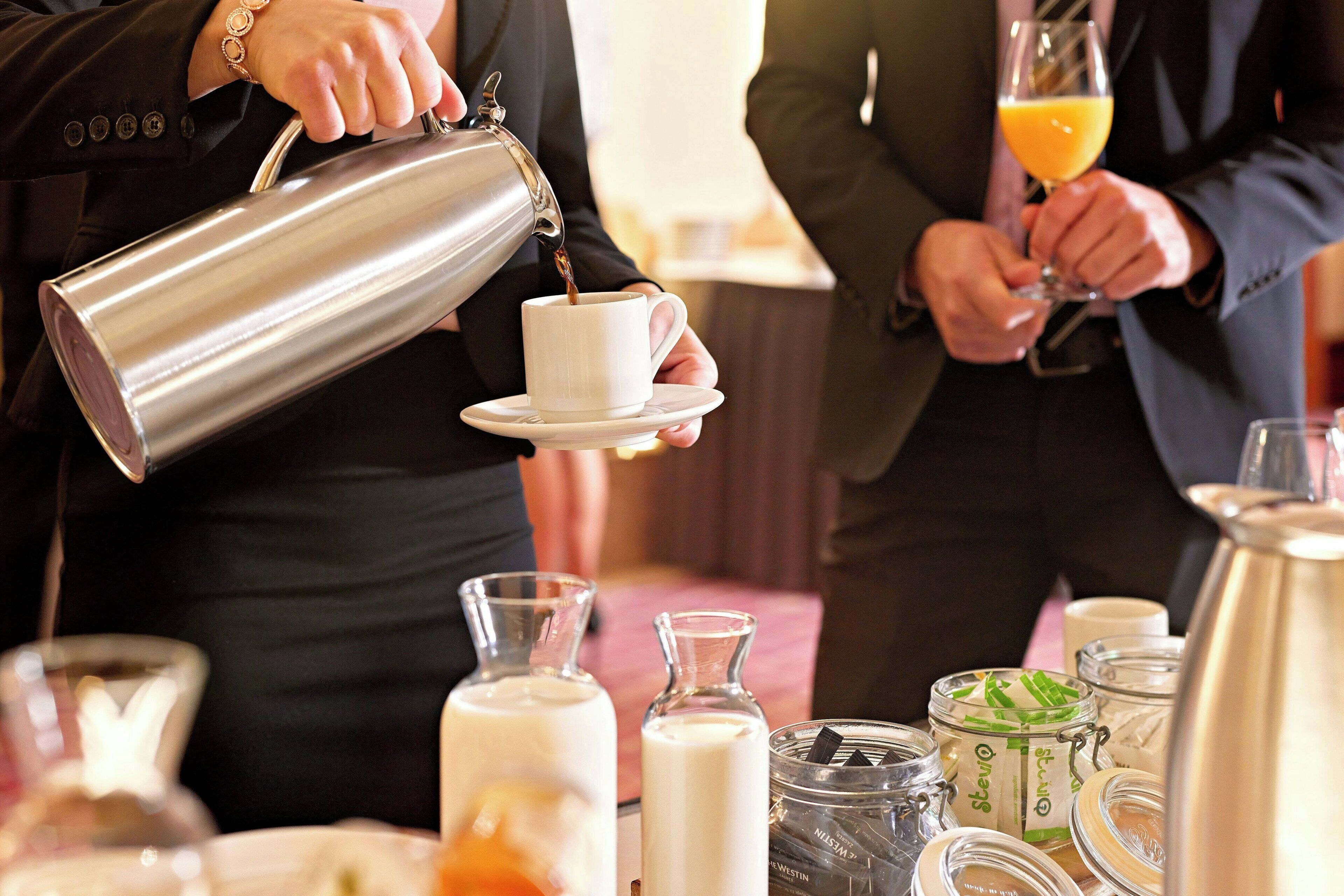 En person hælder kaffe i en kop, et glas appelsinjuice og forskellige beholdere på et bord.