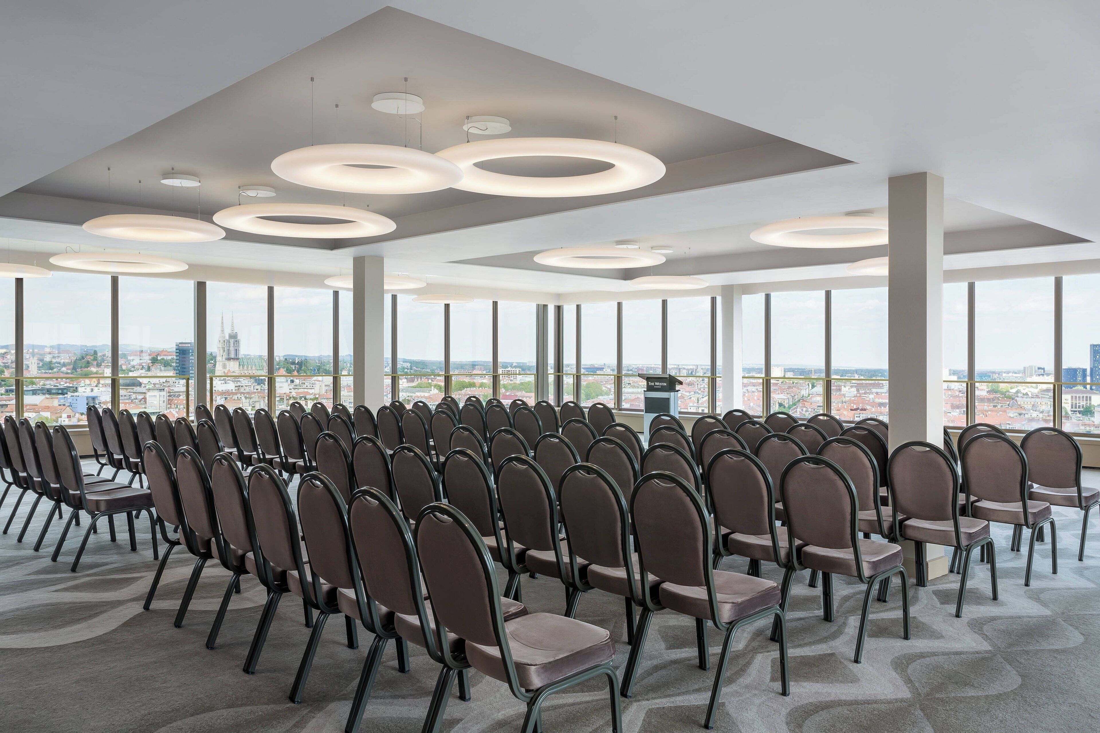 Un salón de conferencias con filas de sillas, ventanas amplias y lámparas circulares en el techo.