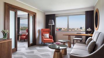 Quarto de hotel moderno com sofá, poltrona e escrivaninha com luminária. Janelas amplas oferecem vista para a cidade.