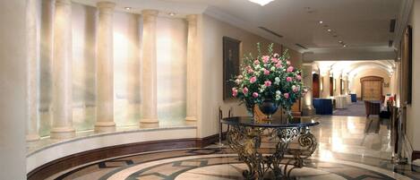 A grand lobby with a marble floor, columns, and a central table with a flower arrangement.