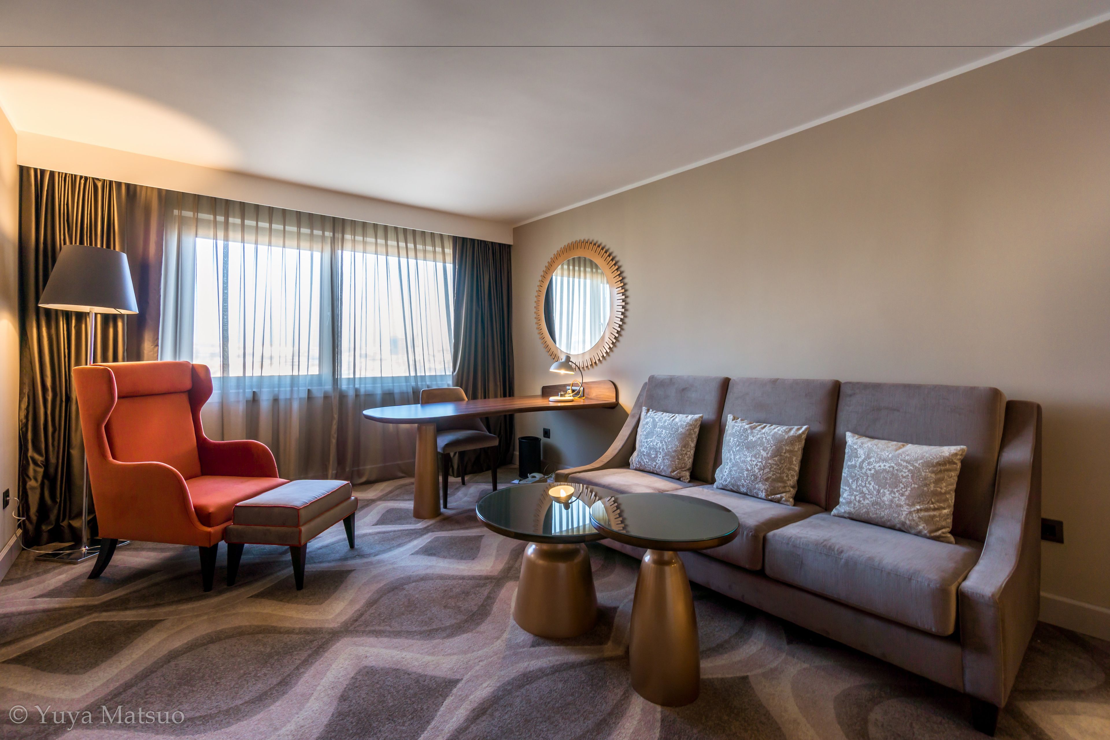 Een moderne hotelkamer met een bank, fauteuil, rond bijzettafeltje en een bureau met lamp.
