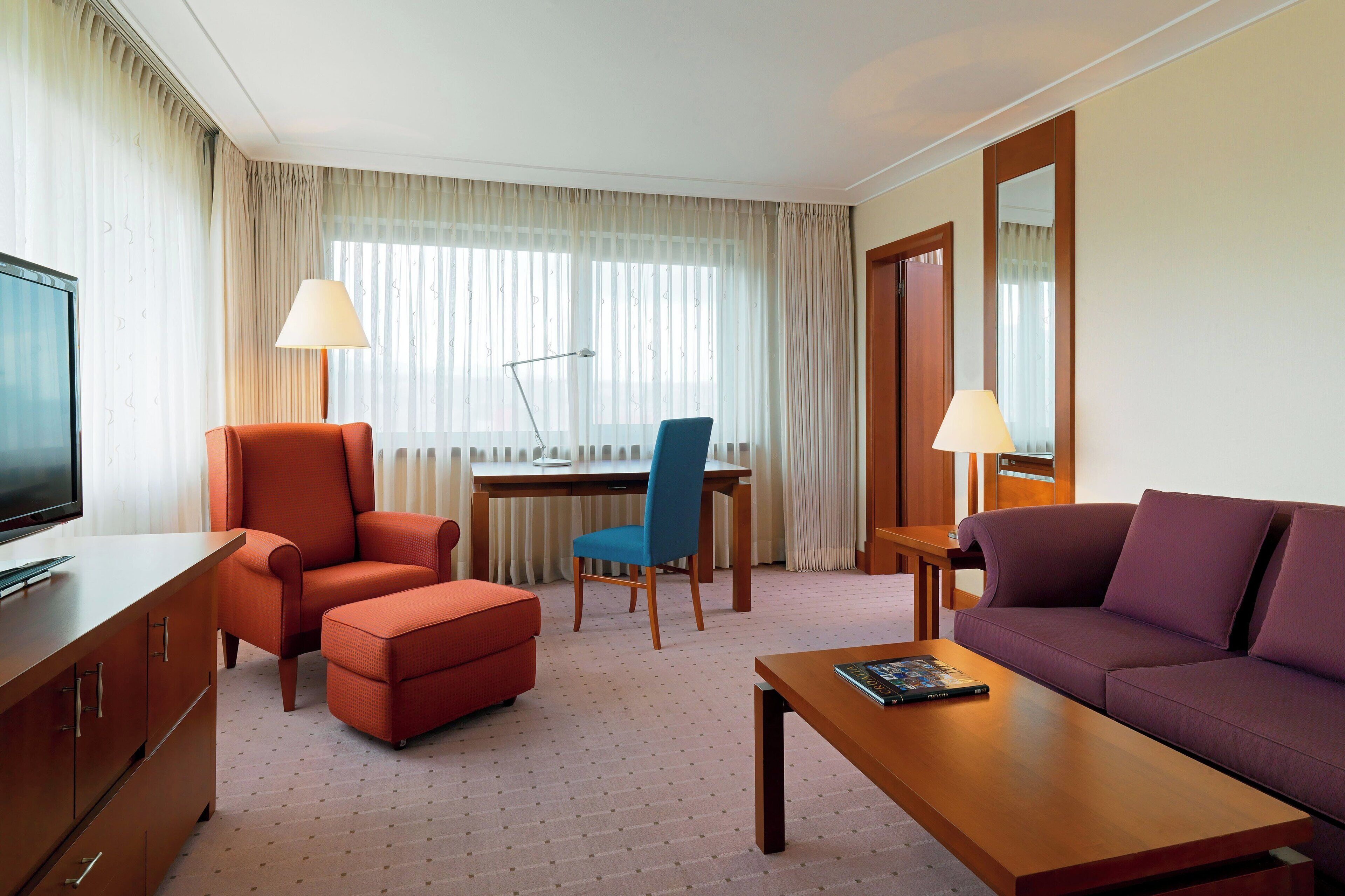 Habitación de hotel con televisor, sillón naranja, silla azul, sofá púrpura, mesita de centro de madera y escritorio con lámparas.