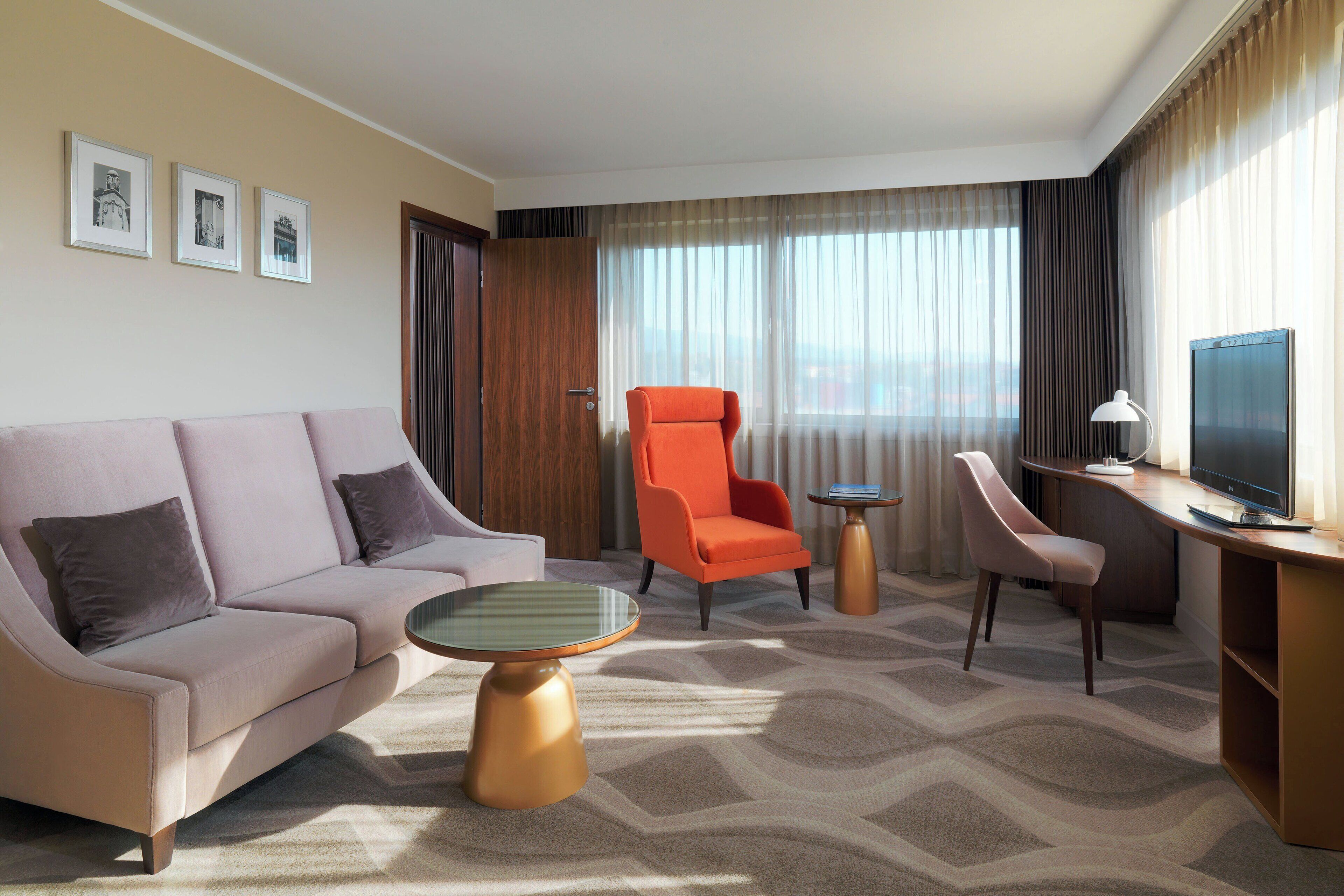 A hotel room with a sofa, an orange armchair, a desk with a TV, and a patterned carpet.