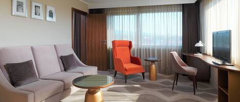A hotel room with a sofa, an orange armchair, a desk with a TV, and a patterned carpet.