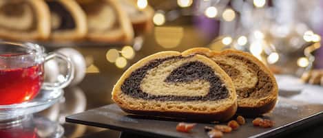 Una rebanada de rollito de chocolate y semillas de amapola sobre un plato negro, junto a un vaso de bebida roja y un fondo borroso de rollitos y luces similares.