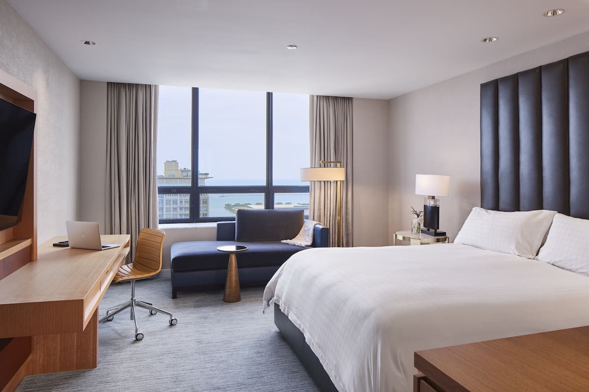 A hotel room with a bed, a desk with a laptop, a chair, a bedside table with a lamp, and a view of the city through large windows.
