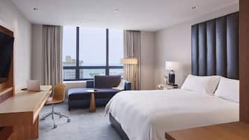A hotel room with a bed, a desk with a laptop, a chair, a bedside table with a lamp, and a view of the city through large windows.