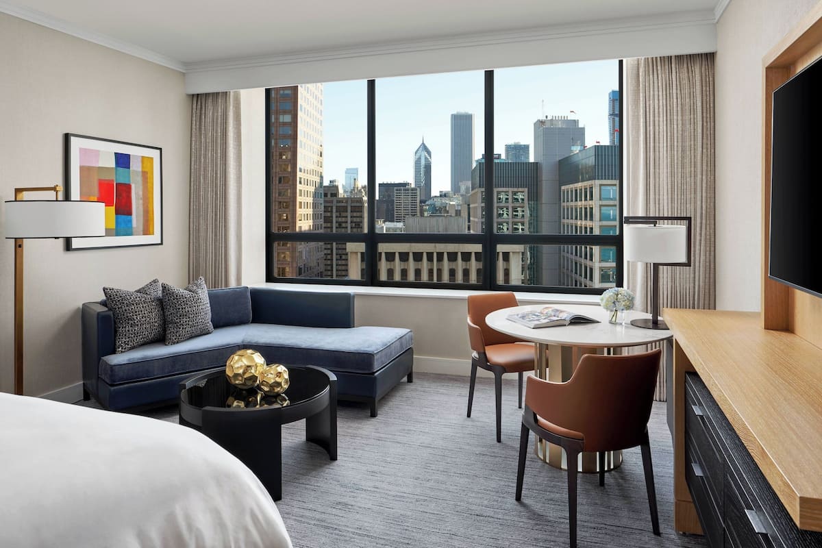 A hotel room with a large window, a sofa, a dining table, and a TV.