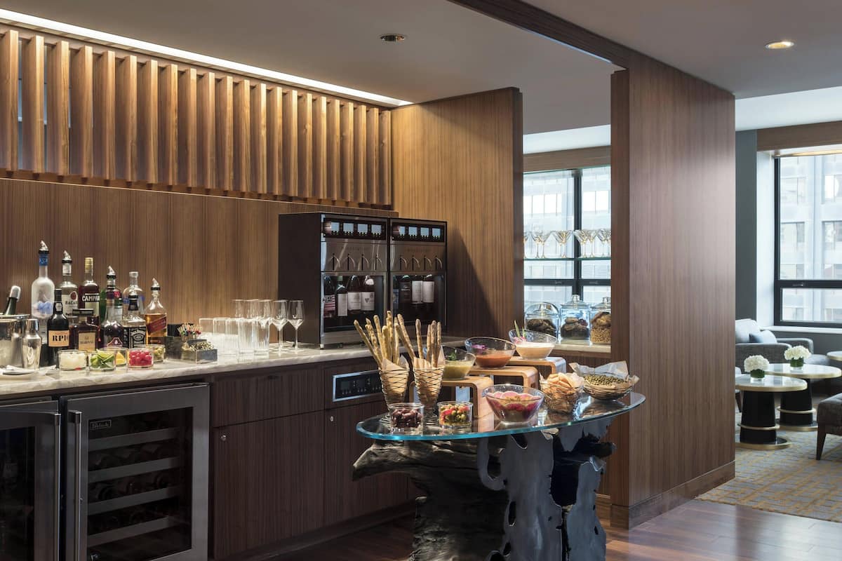 A modern bar area with a wine cooler, bar stools, and a glass-top table with snacks and condiments.