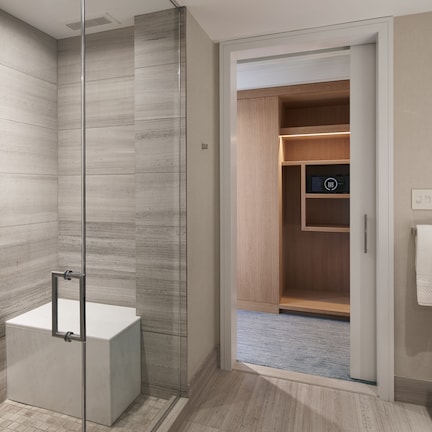 A modern bathroom with a glass-enclosed shower, a white bathtub, and a wooden closet with built-in shelves.