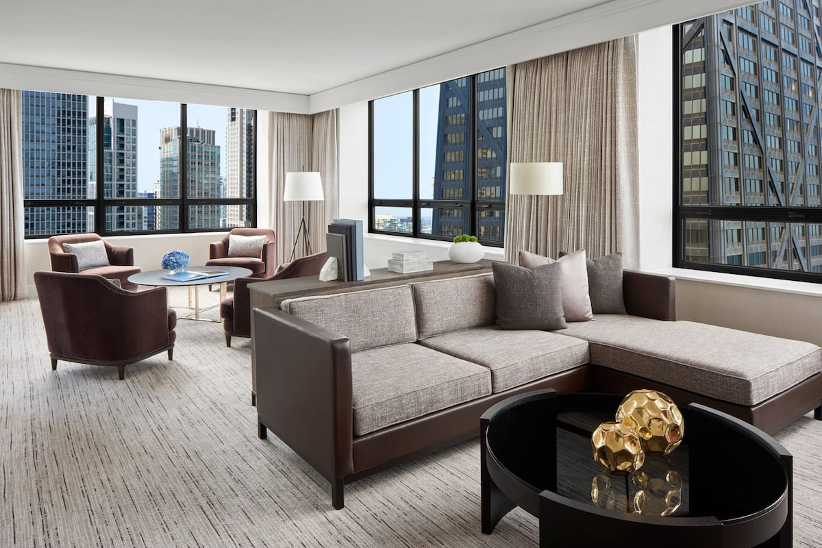 A modern hotel room with a large sectional sofa, a round coffee table, and a view of tall buildings through large windows.