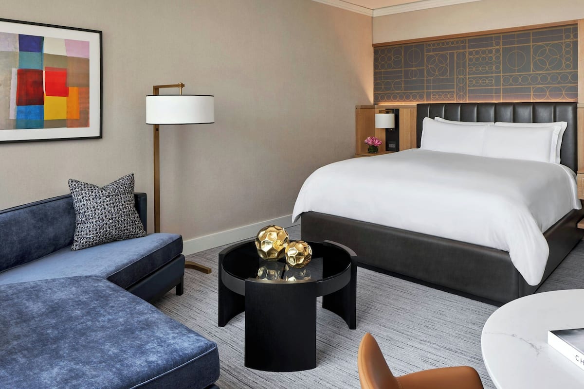 A hotel room with a bed, a sofa, a round black coffee table with decorative items, a lamp, and a painting on the wall.
