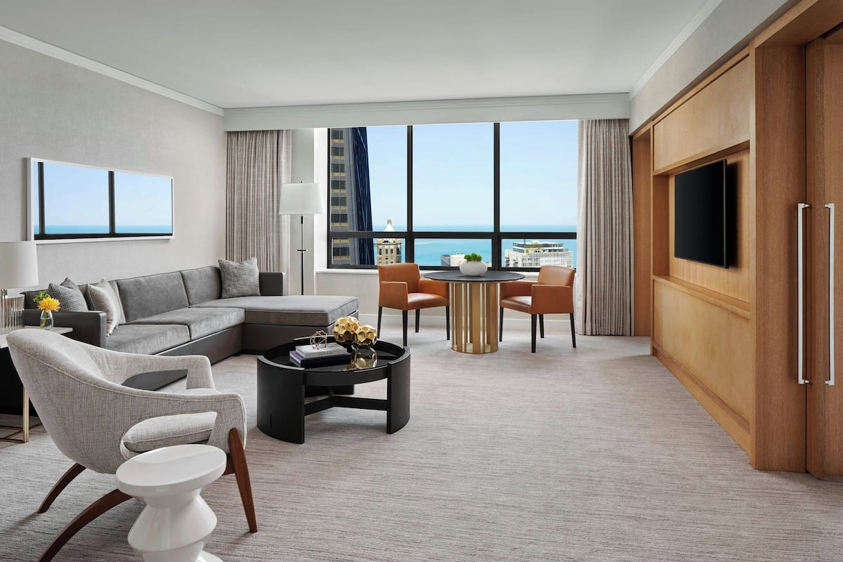 A modern hotel room with a large window offering a view of the city and the sea. It includes a sofa, a dining table, and a TV.